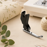 Two black and silver sewing machine presser foot attachments placed on a beige fabric surface, with a sewing machine, thread spool, and fabric roll in the background.